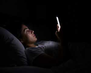 erreurs qui empêchent de bien dormir la nuit: Une personne allongée dans son lit consultant son smartphone lumineux dans une chambre totalement sombre